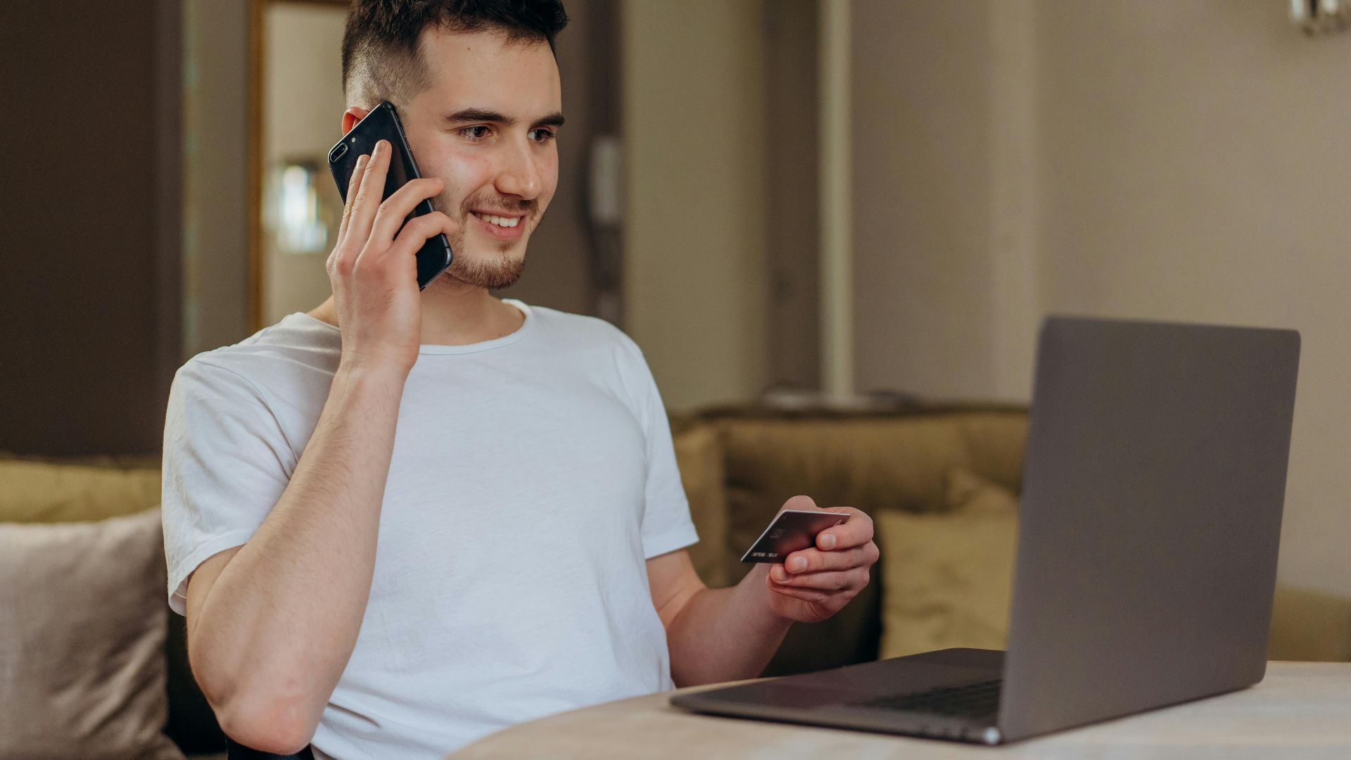 A young man smiles while shopping online using a phone and laptop, holding a credit card.
