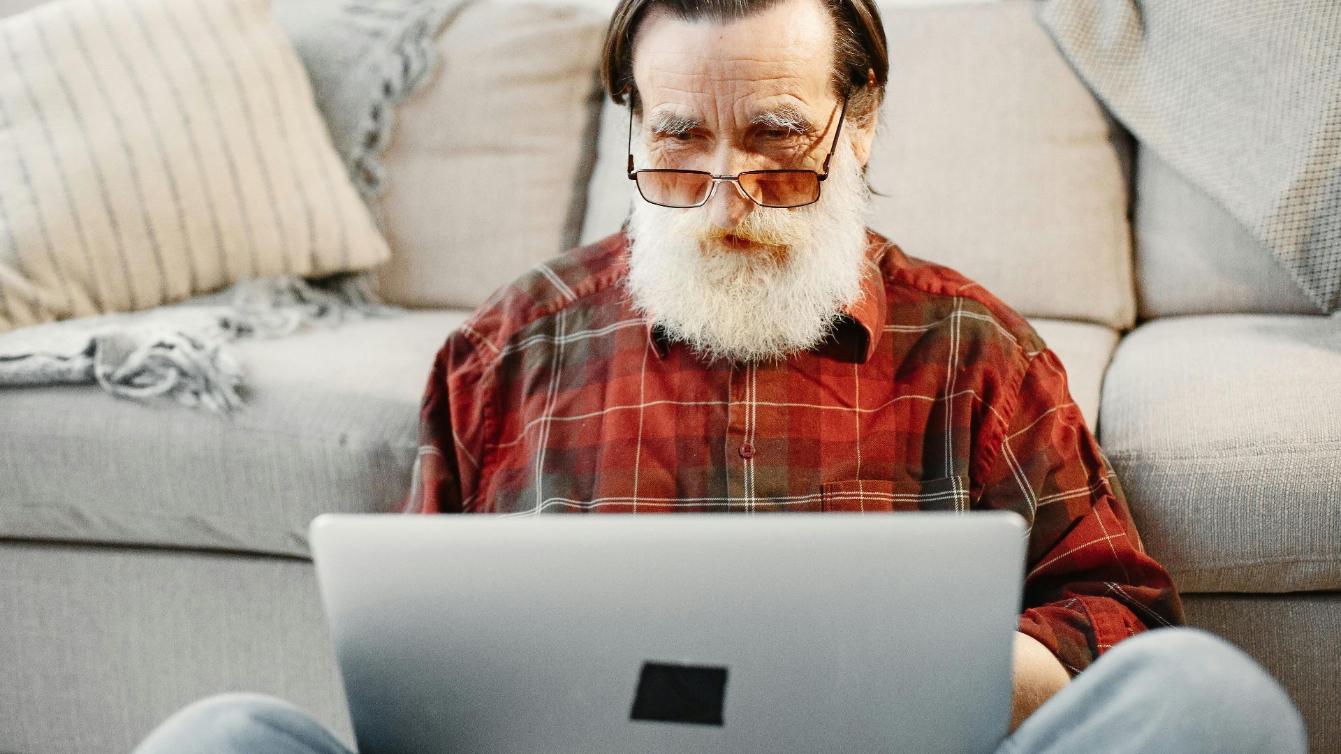 Senior adult man using a laptop while sitting on a couch in a cozy living room setting.