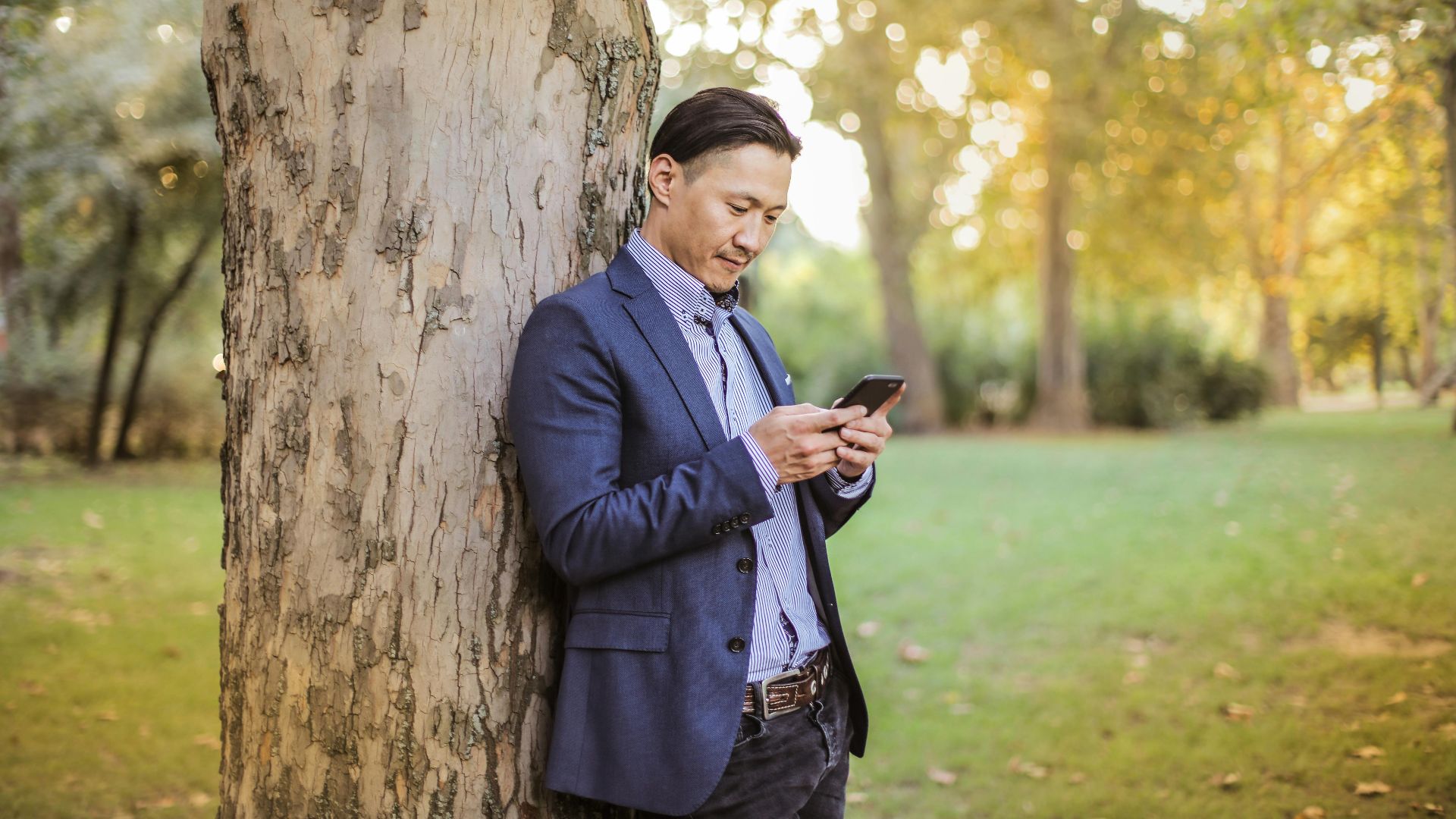 Stylish young man in blue suit using smartphone leaning on tree in a sunny park.
