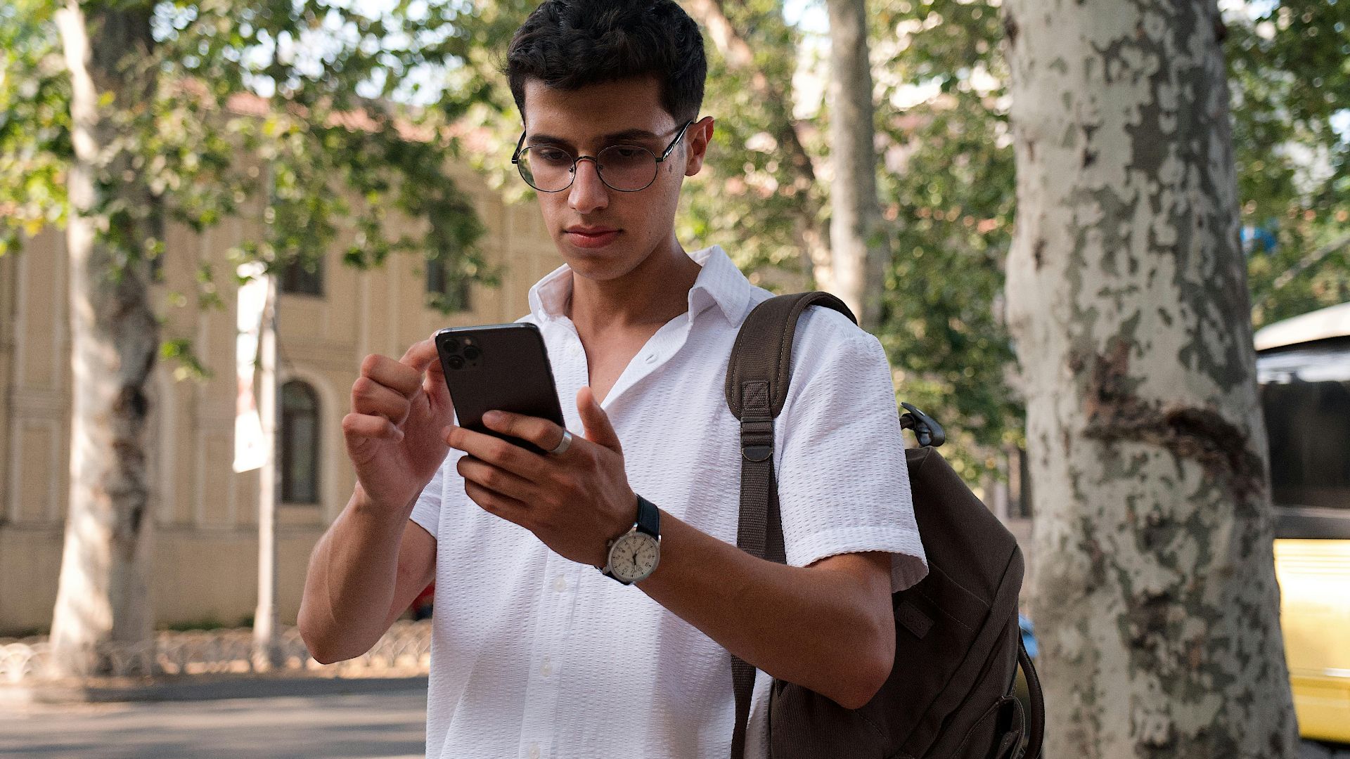Young man using smartphone on a city street, wearing eyeglasses and a backpack.