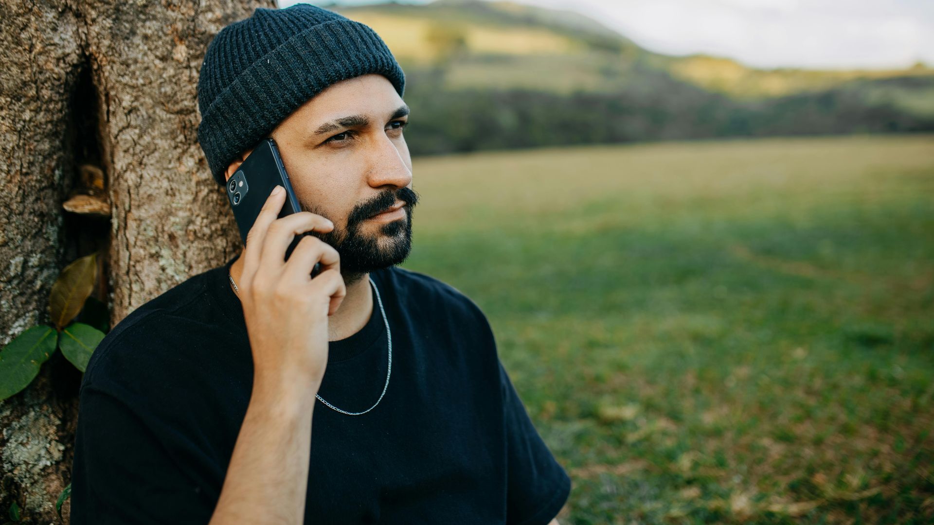 Bearded man wearing hat talking on smartphone while sitting by a tree in a scenic landscape.