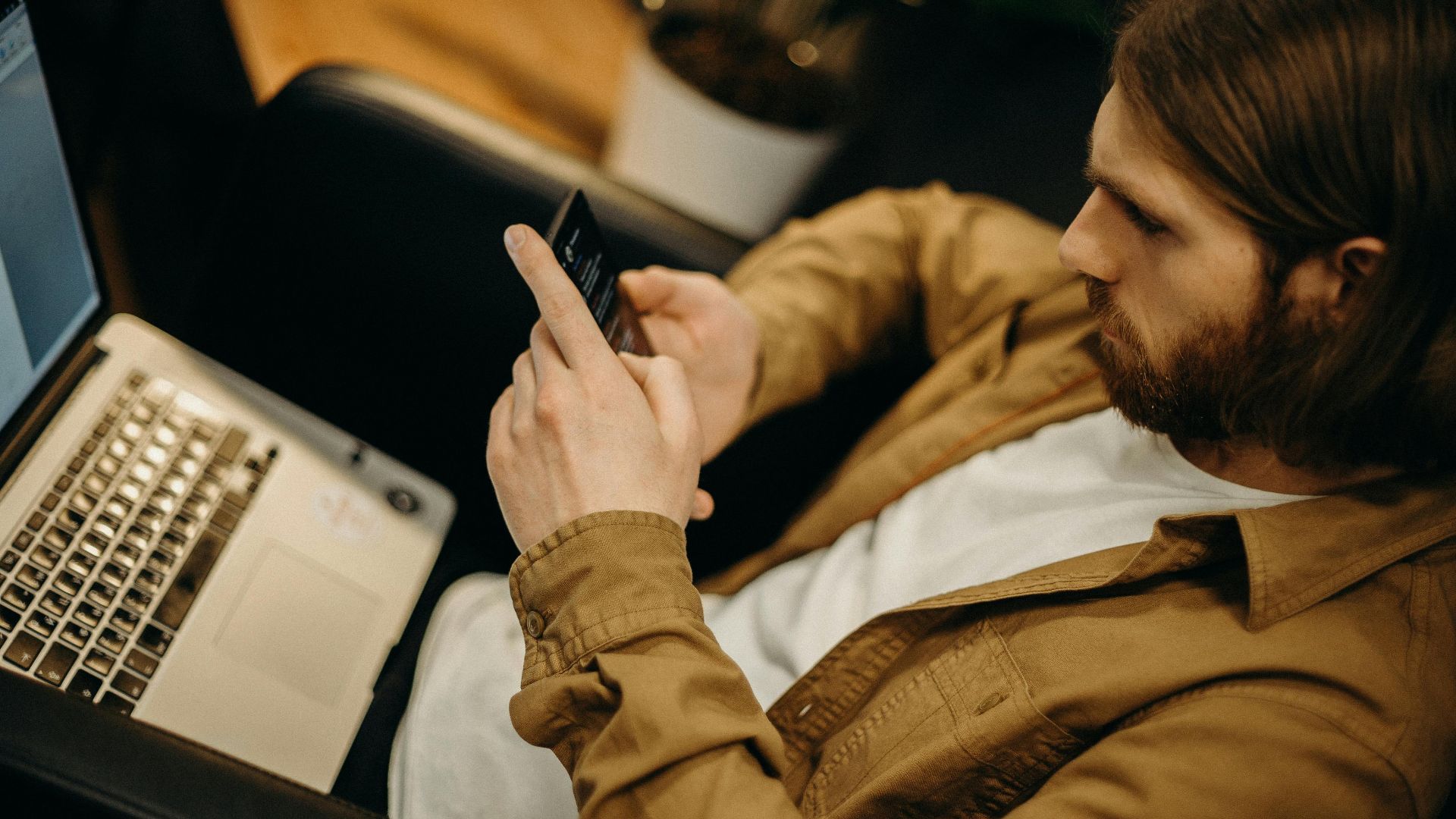 Young man focused on smartphone in a modern workspace with a laptop nearby.
