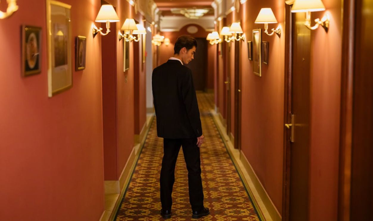 A man in a classic black suit walking through an elegant hotel corridor with warm lighting