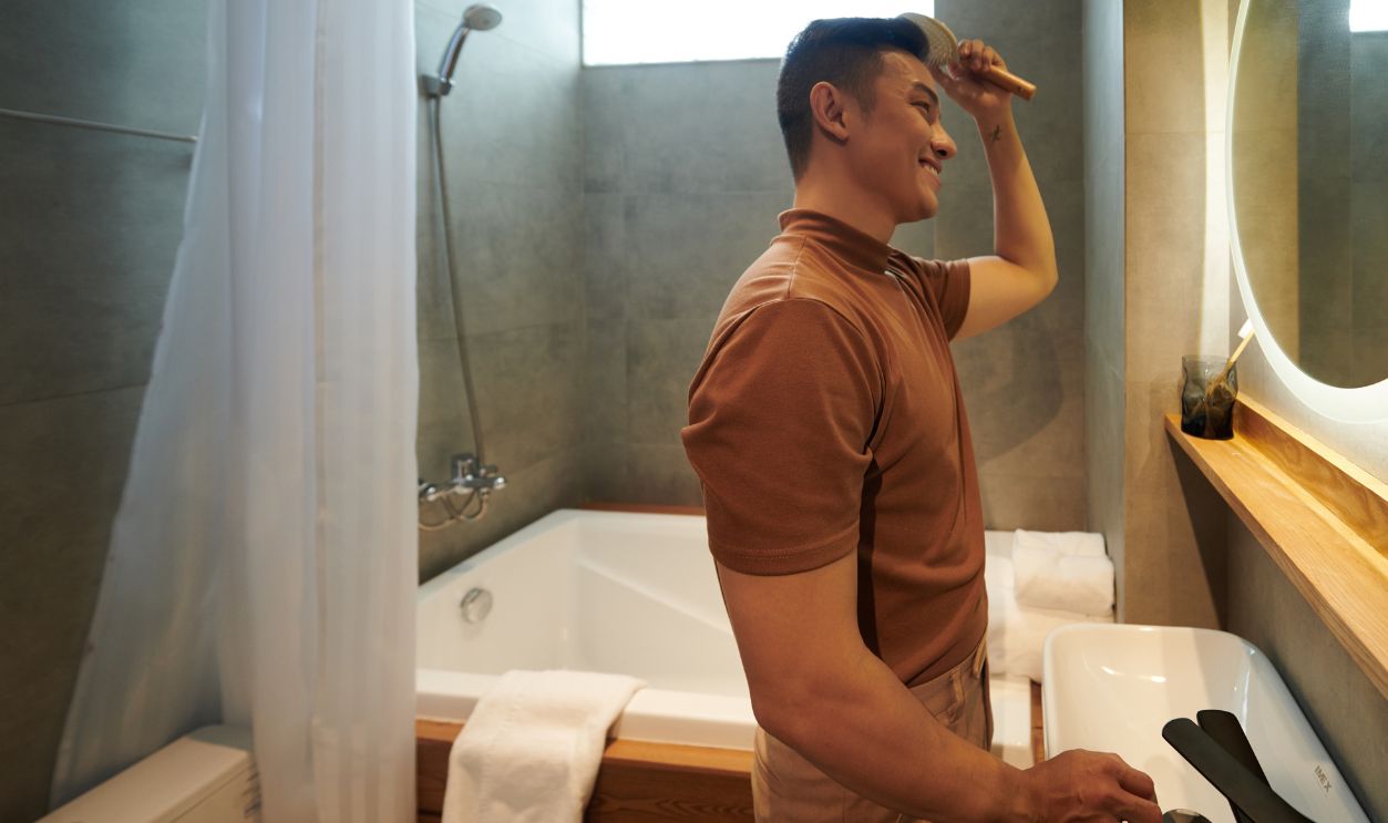 Cheerful Asian young man standing at bathroom window and brushing hair when getting ready in the morning