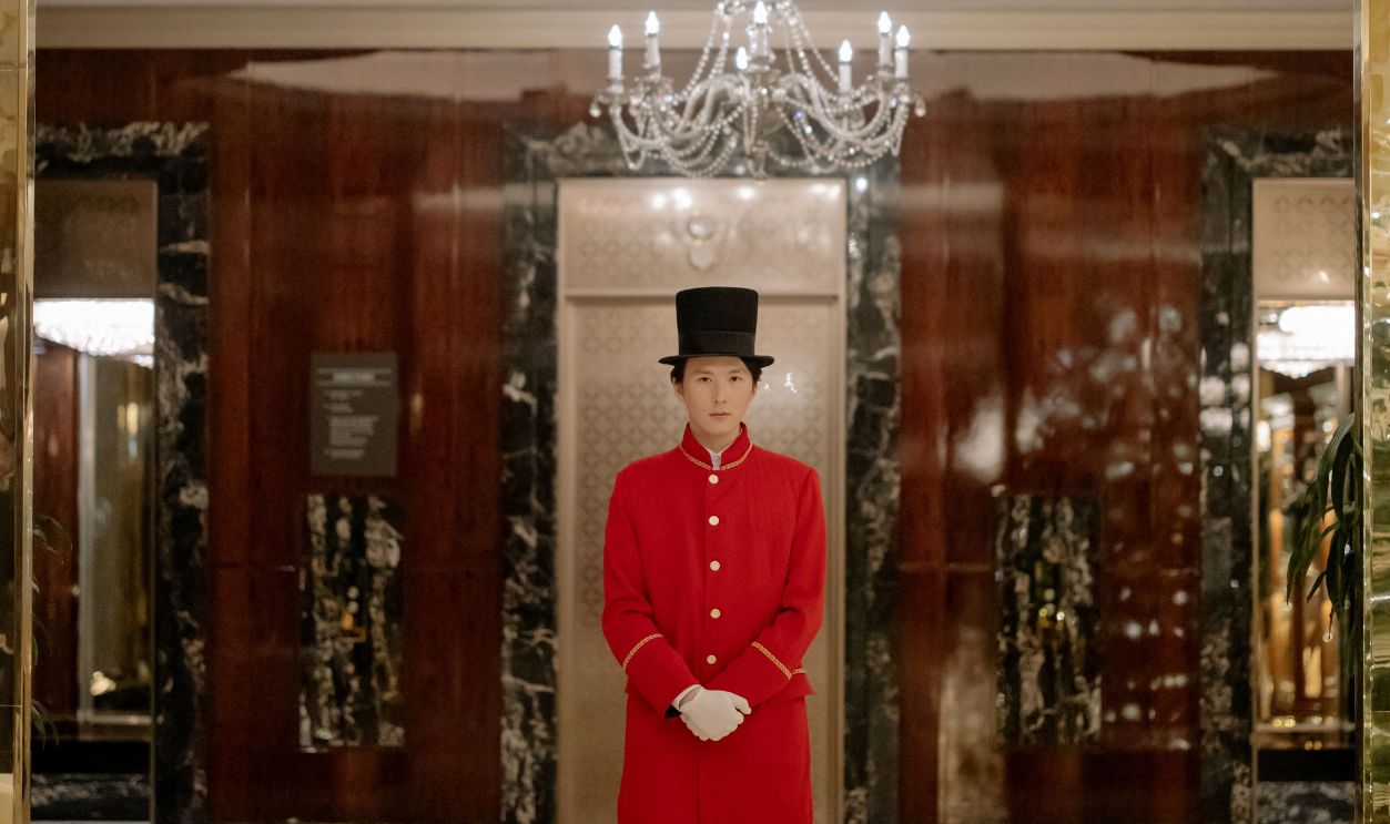 A uniformed concierge in a luxurious hotel lobby with chandeliers and marble decor