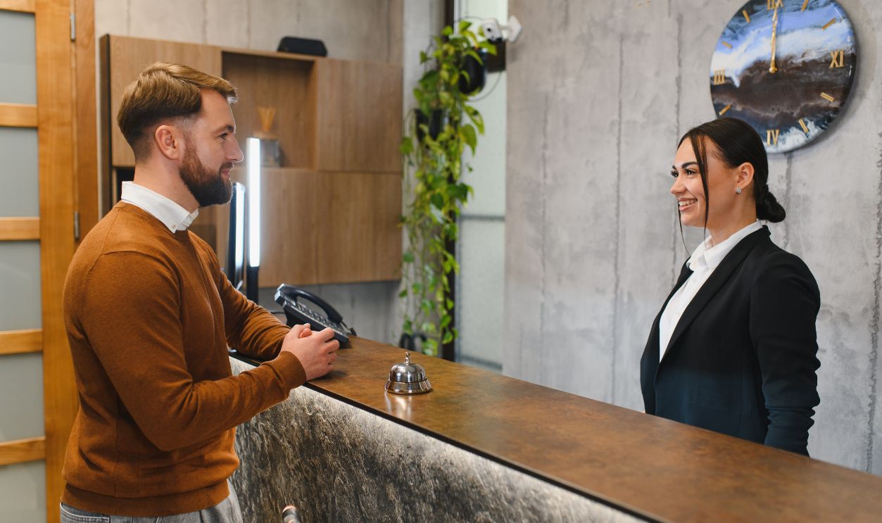 Shutterstock-2727780783, Male customer communicating with smiling female receptionist at modern hotel lobby reception desk, showing hospitality and service