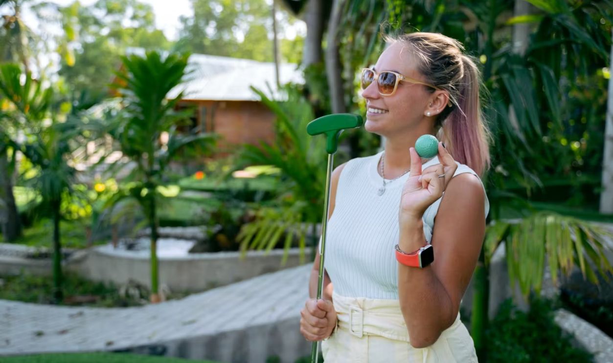 Smiling woman with sunglasses playing mini golf, holding a golf ball and putter outdoors