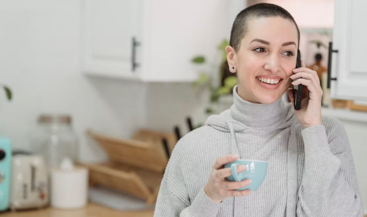 Happy woman with short hair chatting on smartphone while holding a cup in her kitchen.