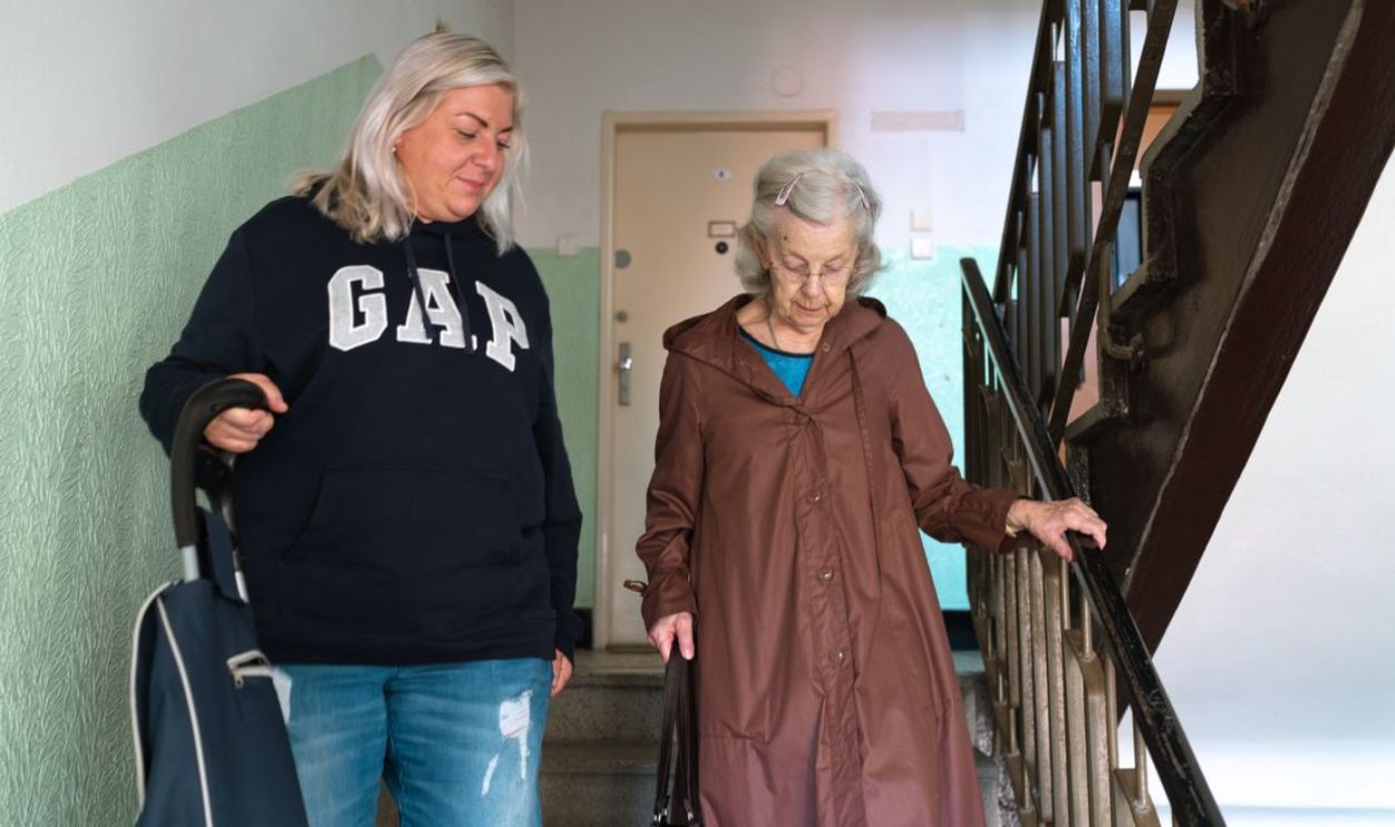 A senior woman assisted by a caregiver walking down indoor stairs in Prague.