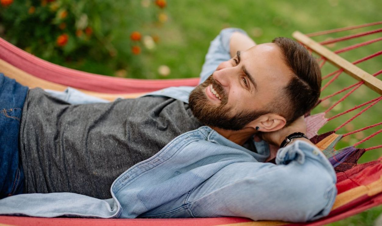 A bearded man in a denim shirt smiles while lying on a colorful hammock outdoors
