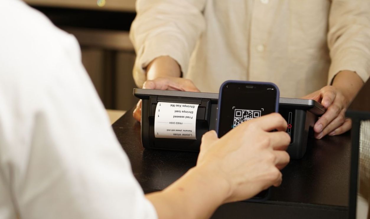 Two individuals engaged in a contactless payment using a QR code scanner at a retail counter.