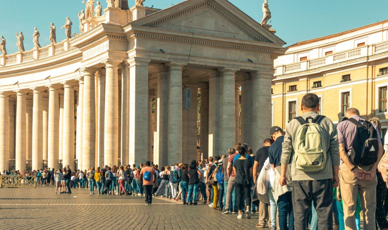 A large crowd of tourists lines up in sunny St. Peter's Square, showcasing architectural columns.