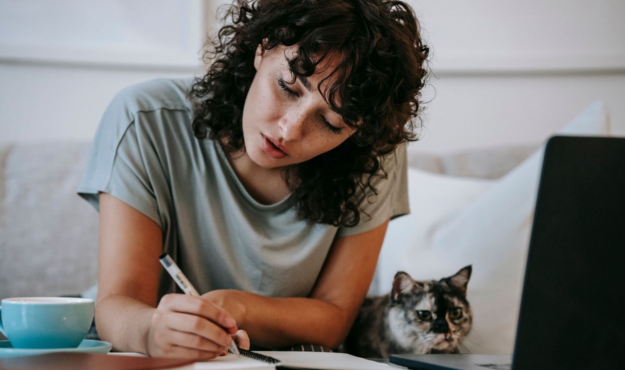 A young woman with curly hair studies at home with her cat nearby