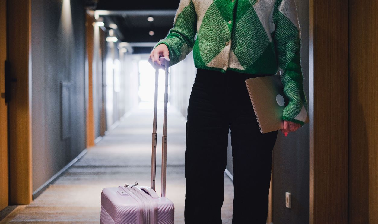 A person in a hotel corridor holding luggage and a laptop