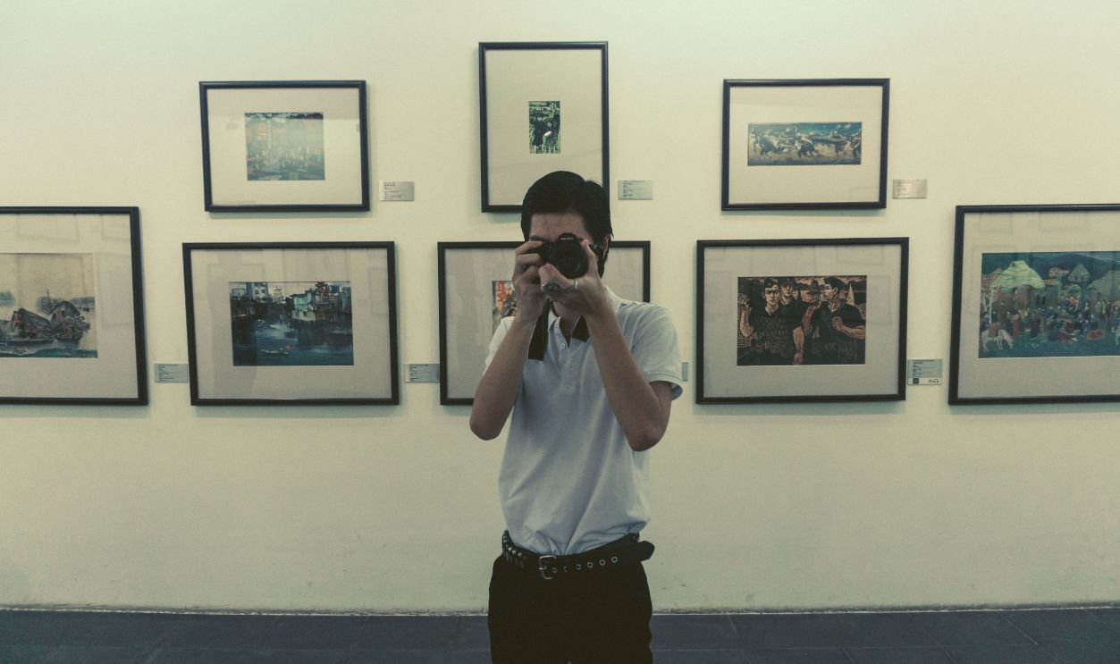 A photographer in a white shirt captures moments in an art gallery filled with framed pictures.