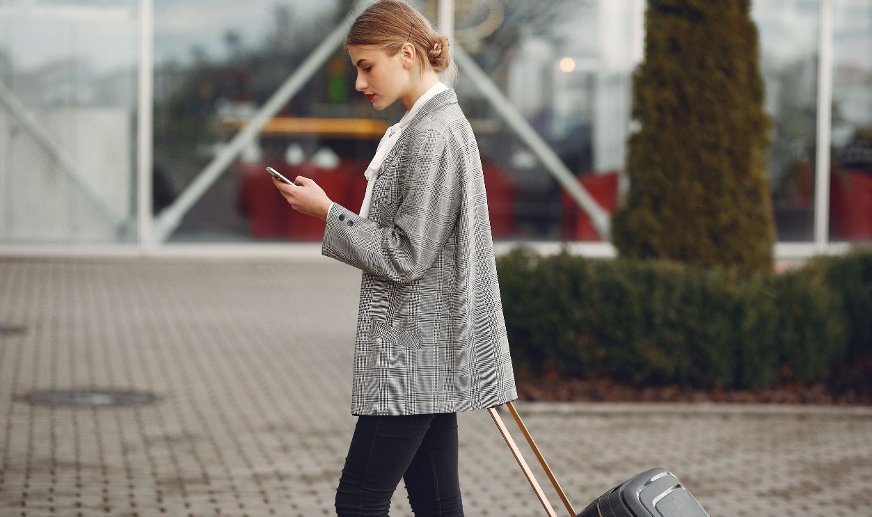Stylish woman walking with suitcase while using smartphone