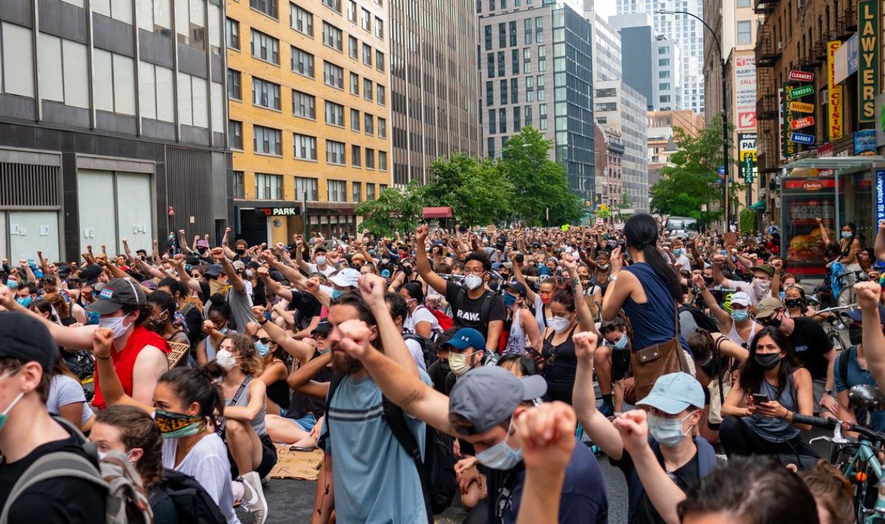 A large crowd of diverse protesters raising fists in a city street during daytime.