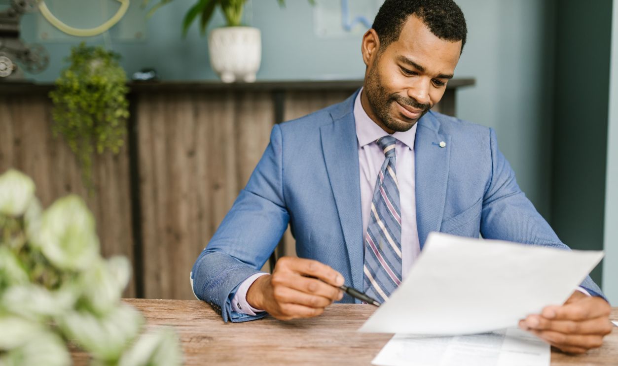 Smiling businessman in blue suit reviewing documents at a wooden table in modern office setting