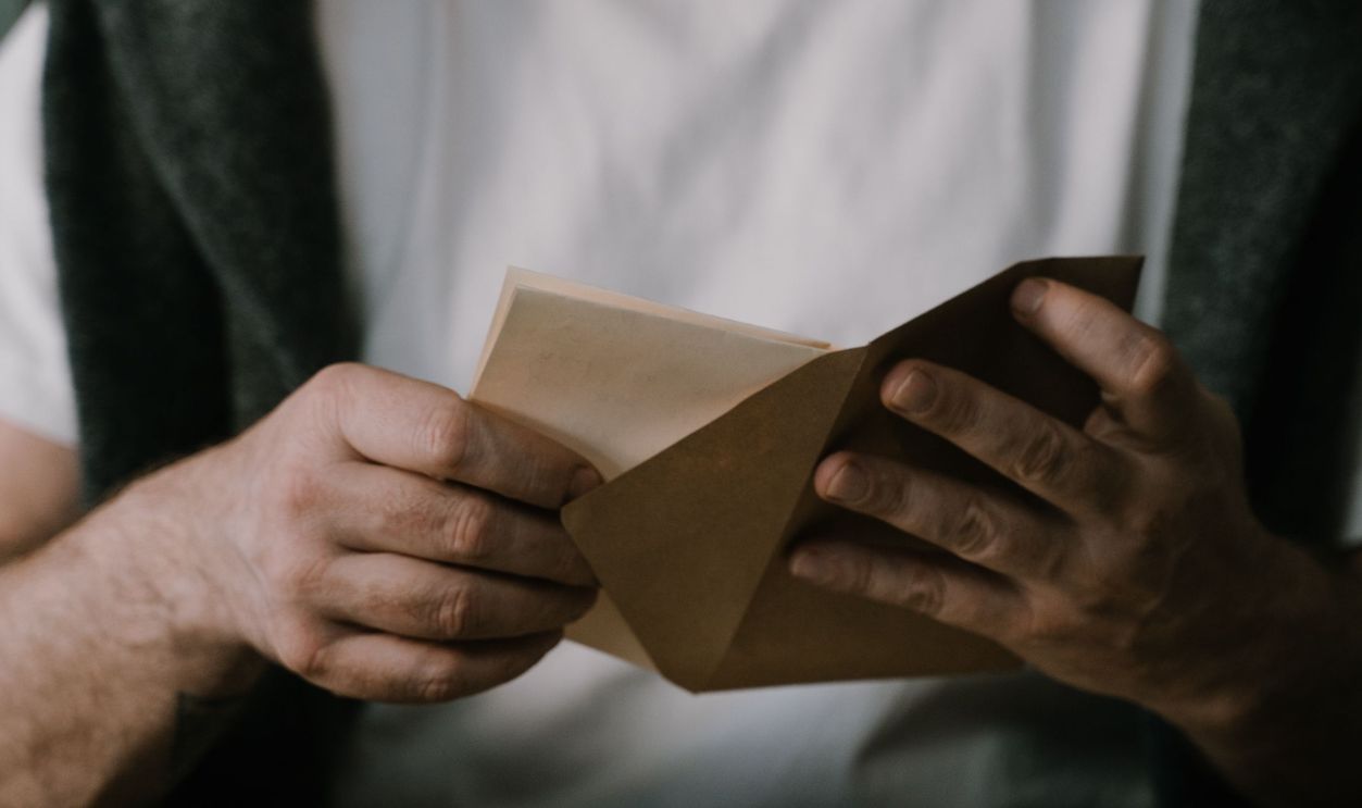 Elderly man with white beard opening an envelope indoors at a rustic table