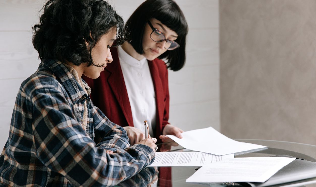 Two women reviewing and signing documents at an indoor glass table