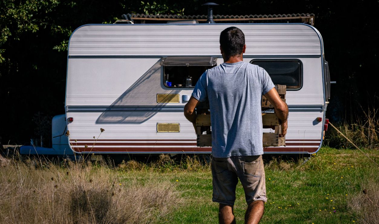 A man carrying a wooden crate approaches a caravan in a sunlit grassy area, evoking a rustic outdoor lifestyle