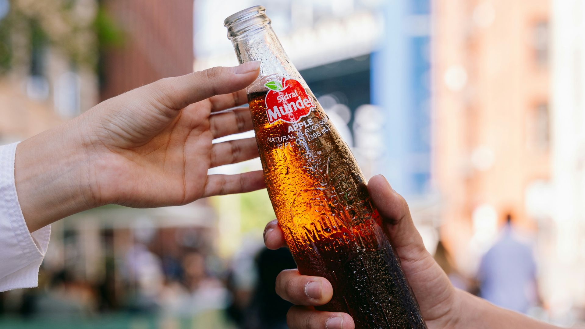 Hands passing a bottled beverage outdoors.