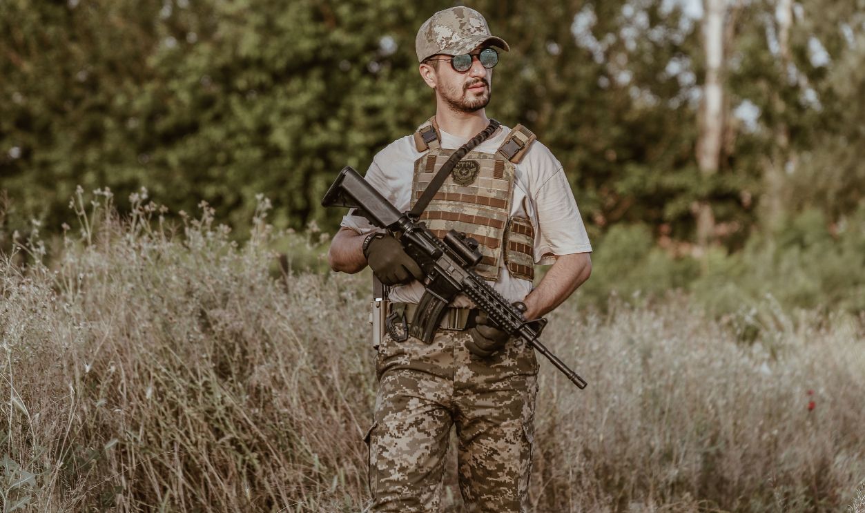 A soldier in camouflage uniform stands with a rifle in a rural grassland area.