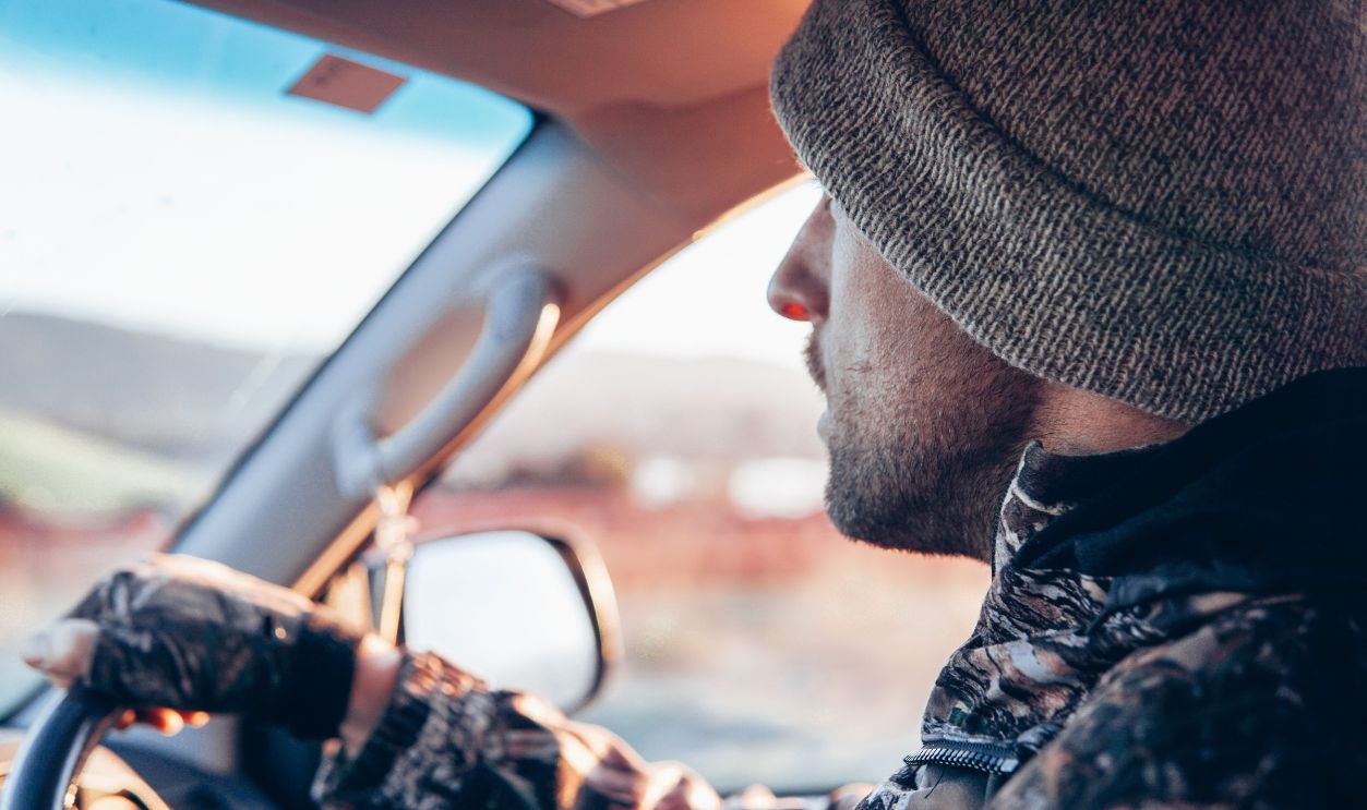 A man wearing a beanie and camouflage jacket drives a car at sunrise, capturing the essence of freedom and adventure.