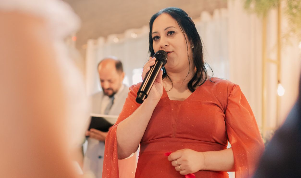 Woman in red dress speaking at indoor ceremony with microphone