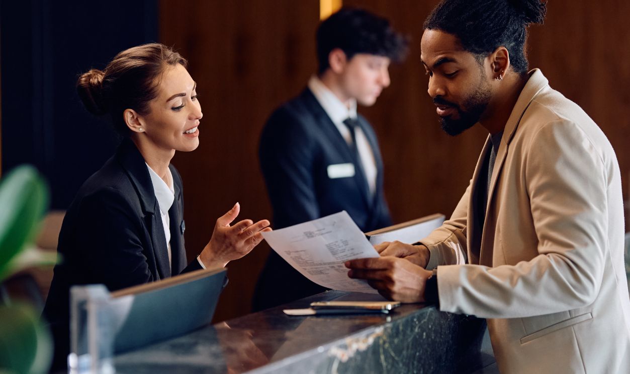 Shutterstock-2441452197, Smiling hotel manager assisting African American guest in filling paperwork at reception desk.