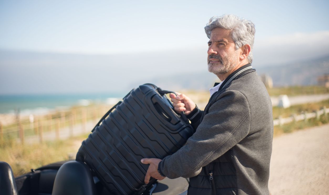Elderly man with suitcase by a convertible on a scenic coastal journey in Portugal.