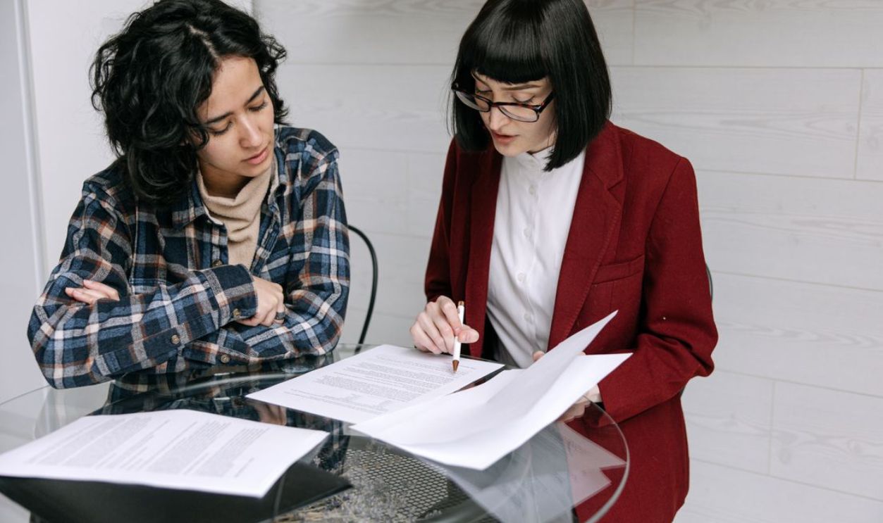 Two women collaborating at a glass table, reviewing business documents in a modern office setting
