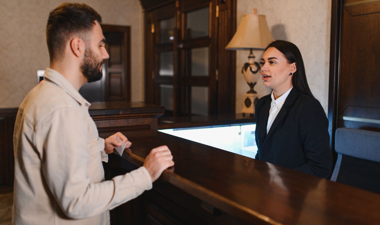 Shutterstock-2714036827, Guest standing at the hotel reception desk, talking to a female receptionist during a vacation or business trip