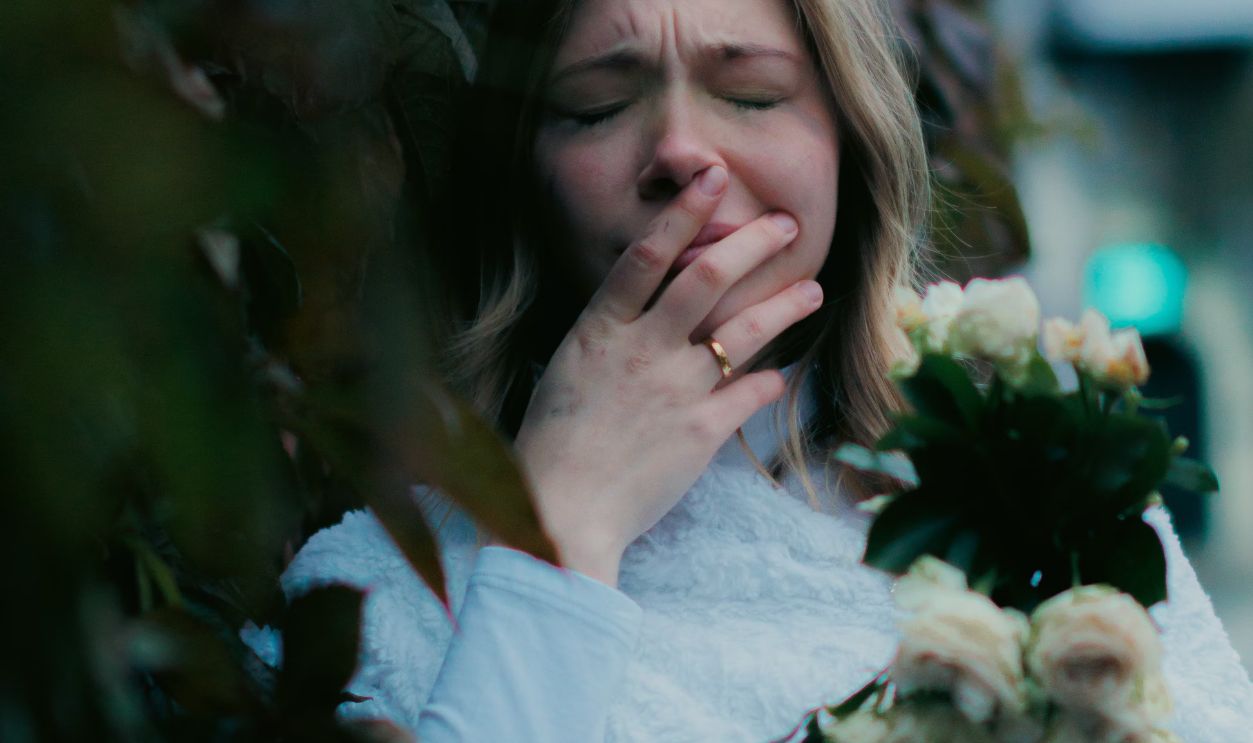 A young woman in a white dress holds a bouquet, leaning against a hedge, eyes closed