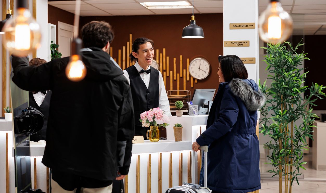 Shutterstock-2482489193, Young couple with winter clothes and suitcase arriving at front desk of luxury holiday resort for room reservation. Friendly concierge welcomes two tourists with ski attire at hotel reception counter.