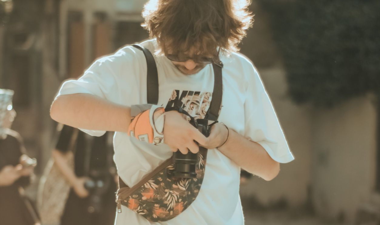 A young man in a white shirt and shorts adjusts his floral belt bag while standing outdoors.