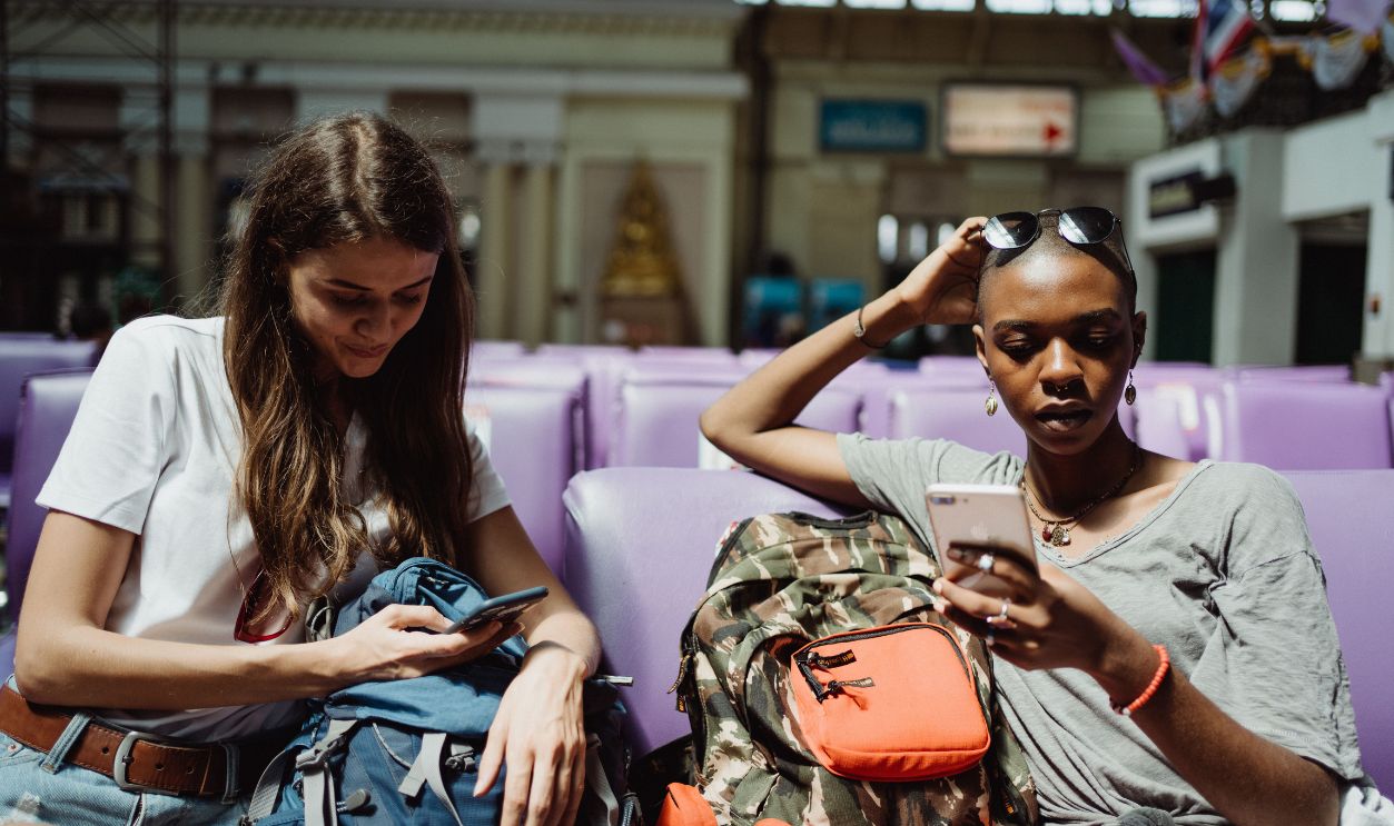 Two women with backpacks using smartphones while waiting