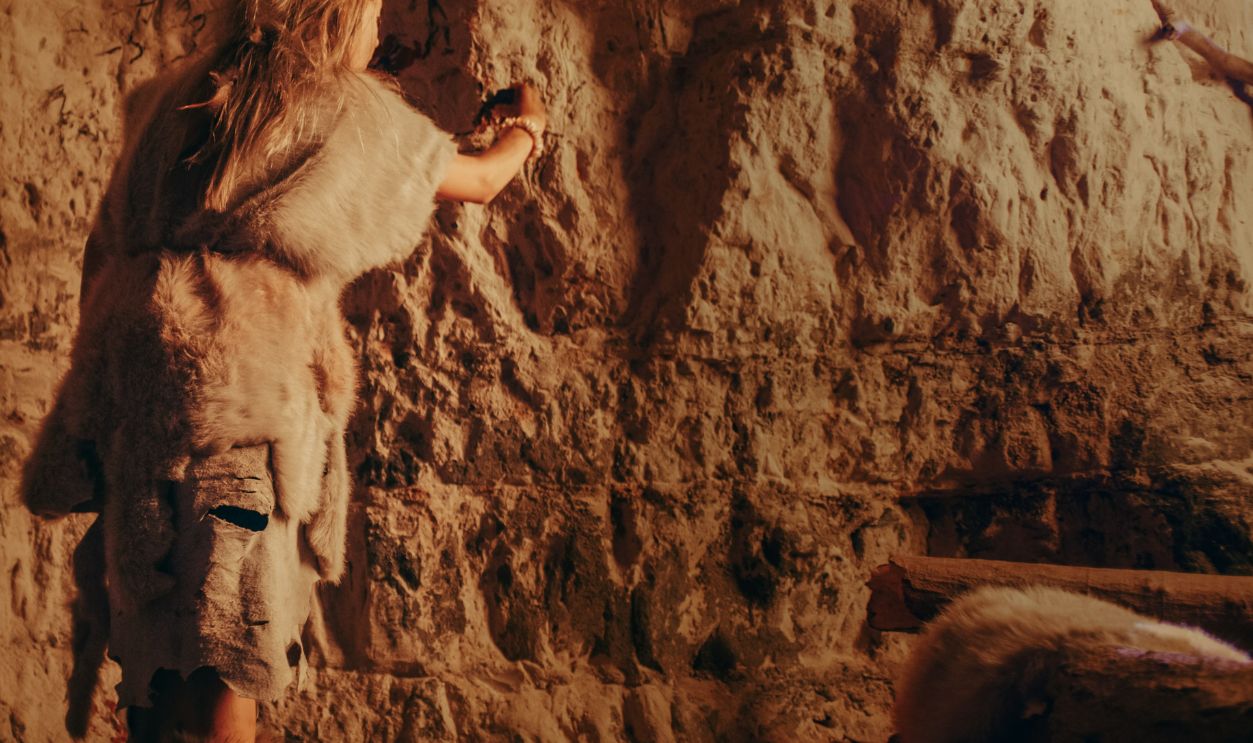 Back View of a Primitive Prehistoric Neanderthal Child Wearing Animal Skin Draws Animals and Abstracts on the Walls at Night. Creating First Cave Art with Petroglyphs, Rock Paintings.