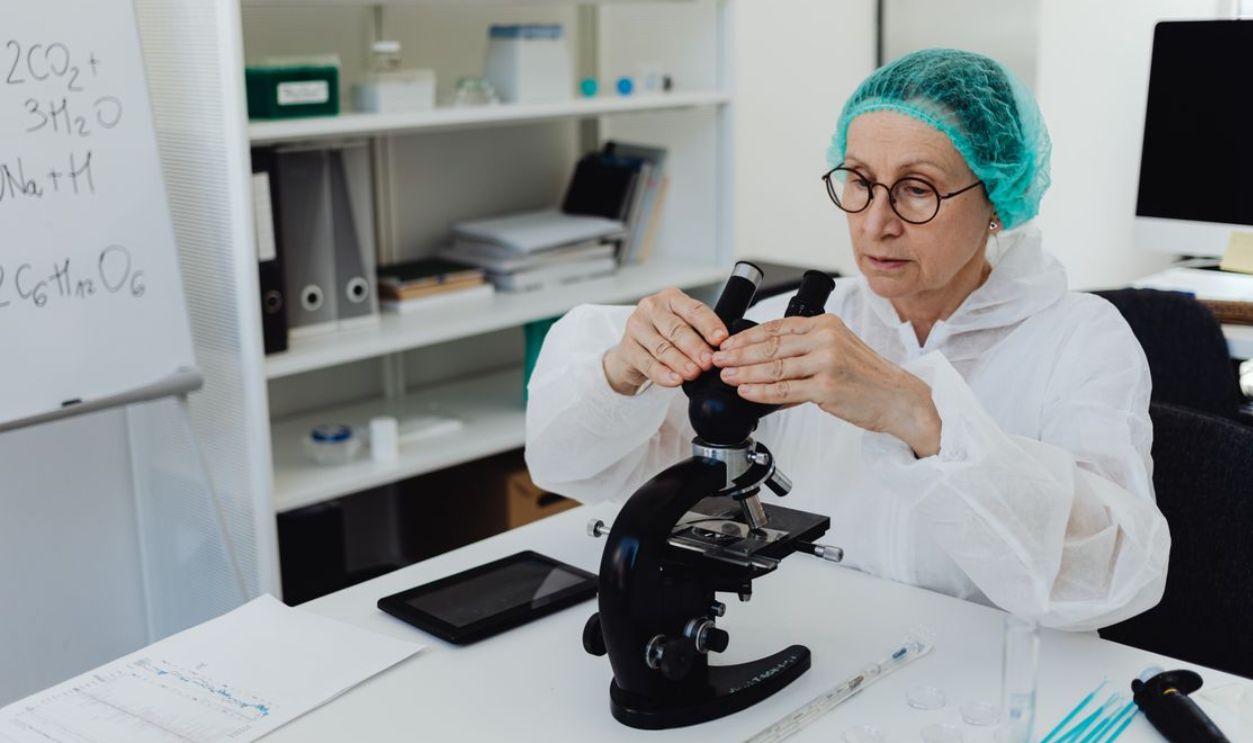 Elderly female scientist in PPE examining samples with a microscope in a modern lab
