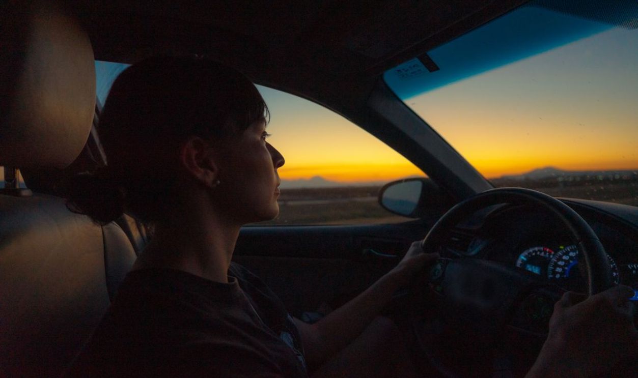 Silhouette of a woman driving a car at sunset with scenic view outside.