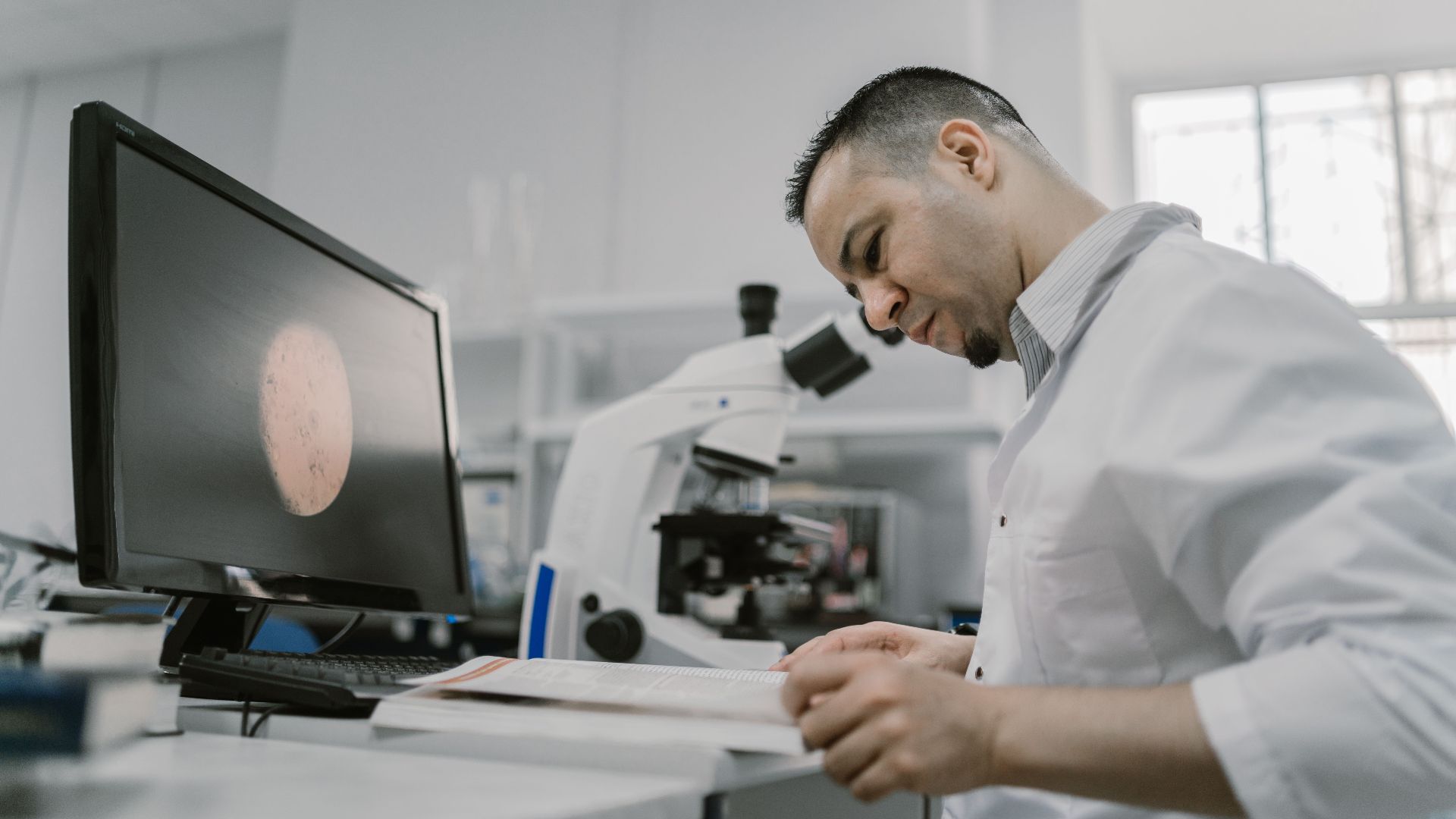 Asian man reading in a laboratory, surrounded by equipment and microscope, focused on work.
