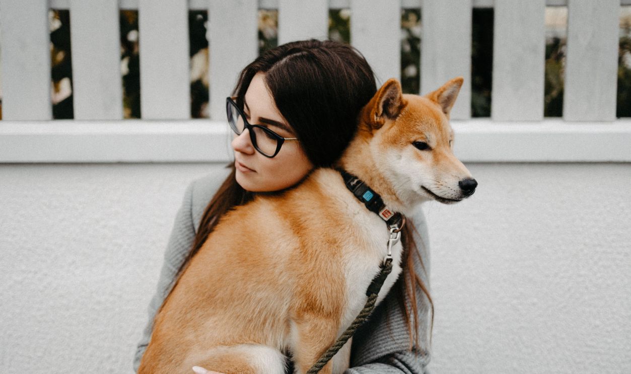 A young woman with glasses cuddles a Shiba Inu dog outdoors