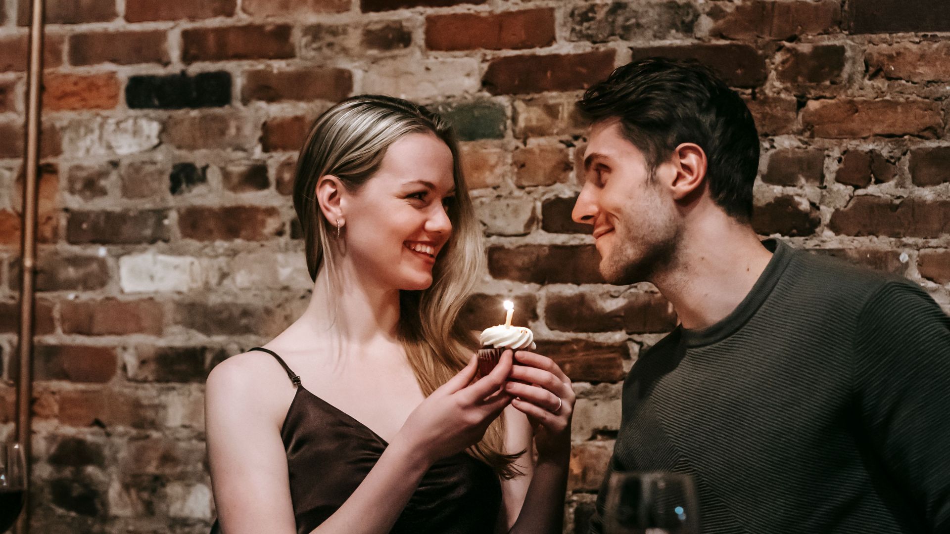 Side view of loving couple sitting near brick wall at wooden table with cupcake with burning candle looking at each other while celebrating anniversary in cafe