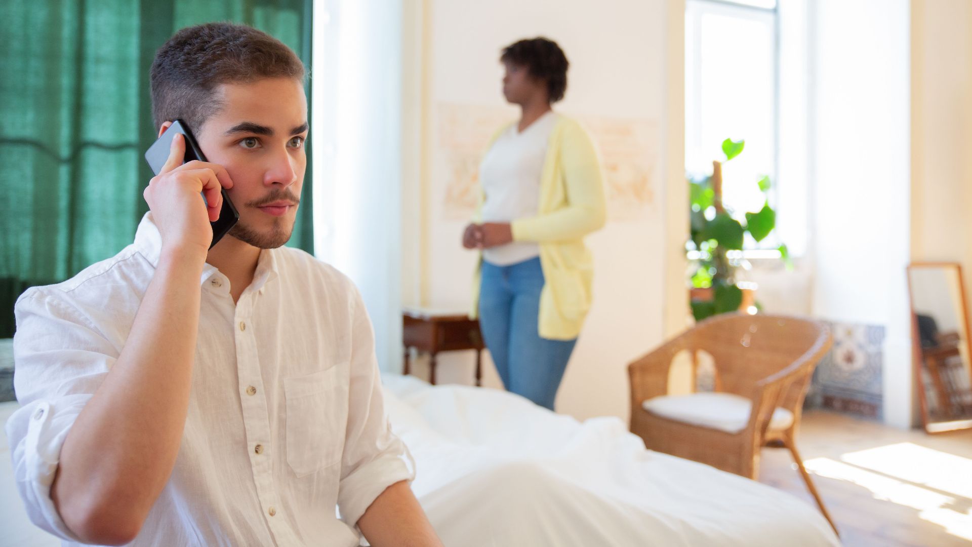 Young man using a smartphone indoors, with a woman standing in the background by the window.
