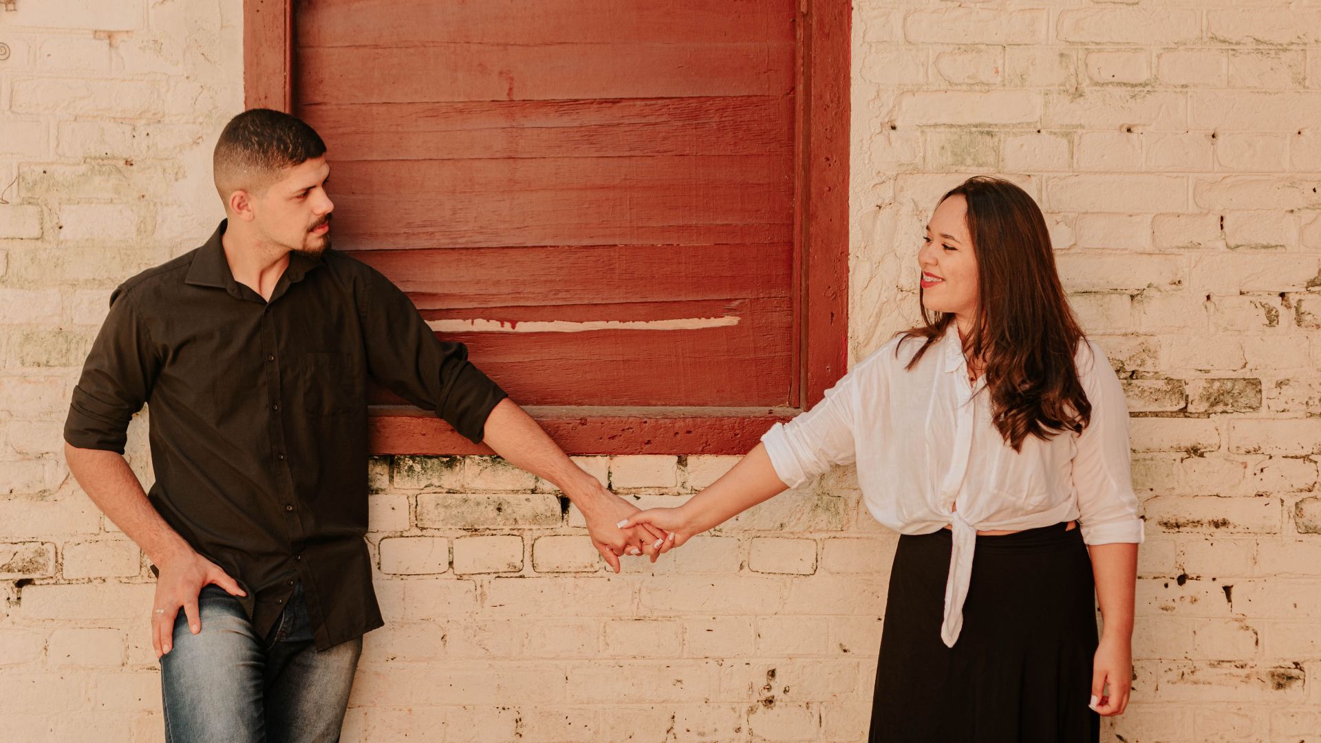 A romantic couple smiling and holding hands against a rustic brick wall, exuding warmth and connection.