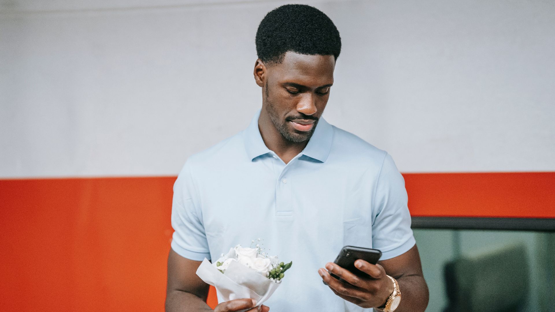 A man in a polo shirt holds flowers and checks his mobile phone.