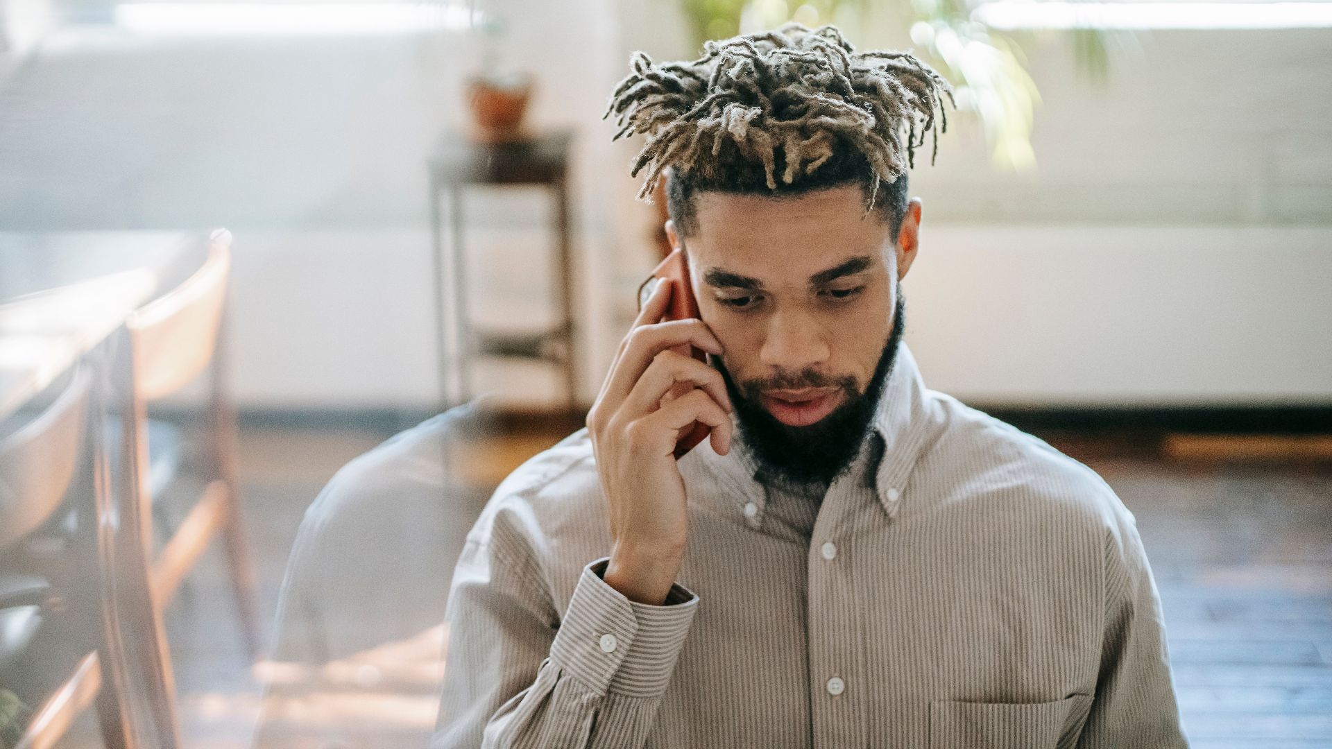 Black man in casual outfit speaking on cellphone in light room near table with window and plant on background