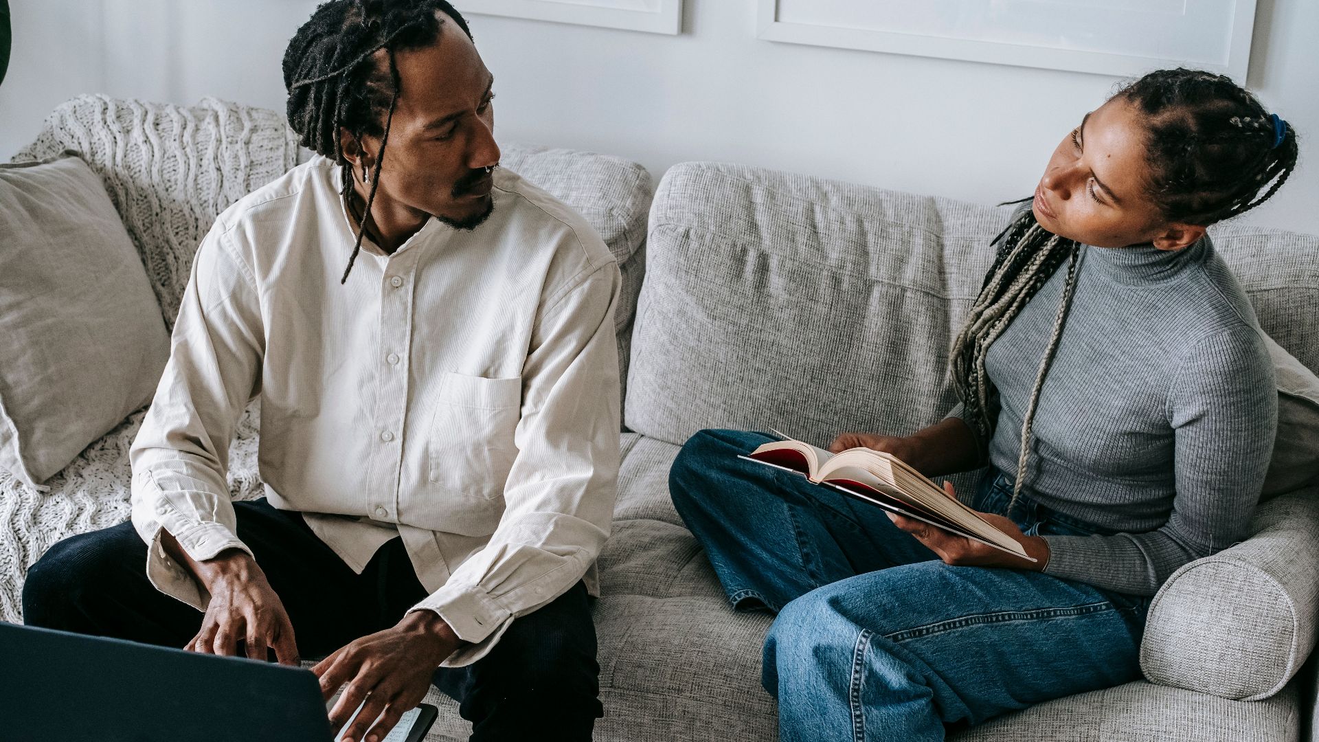 From above of serious young African American husband and wife in casual outfits sitting on sofa with book and laptop and discussing relationships in cozy living room