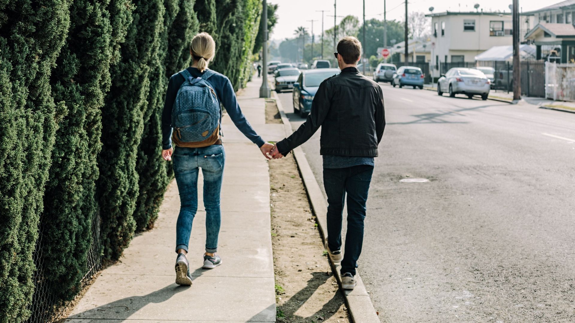 A couple walks hand in hand along a sunny sidewalk in an urban area, showcasing romance and companionship.