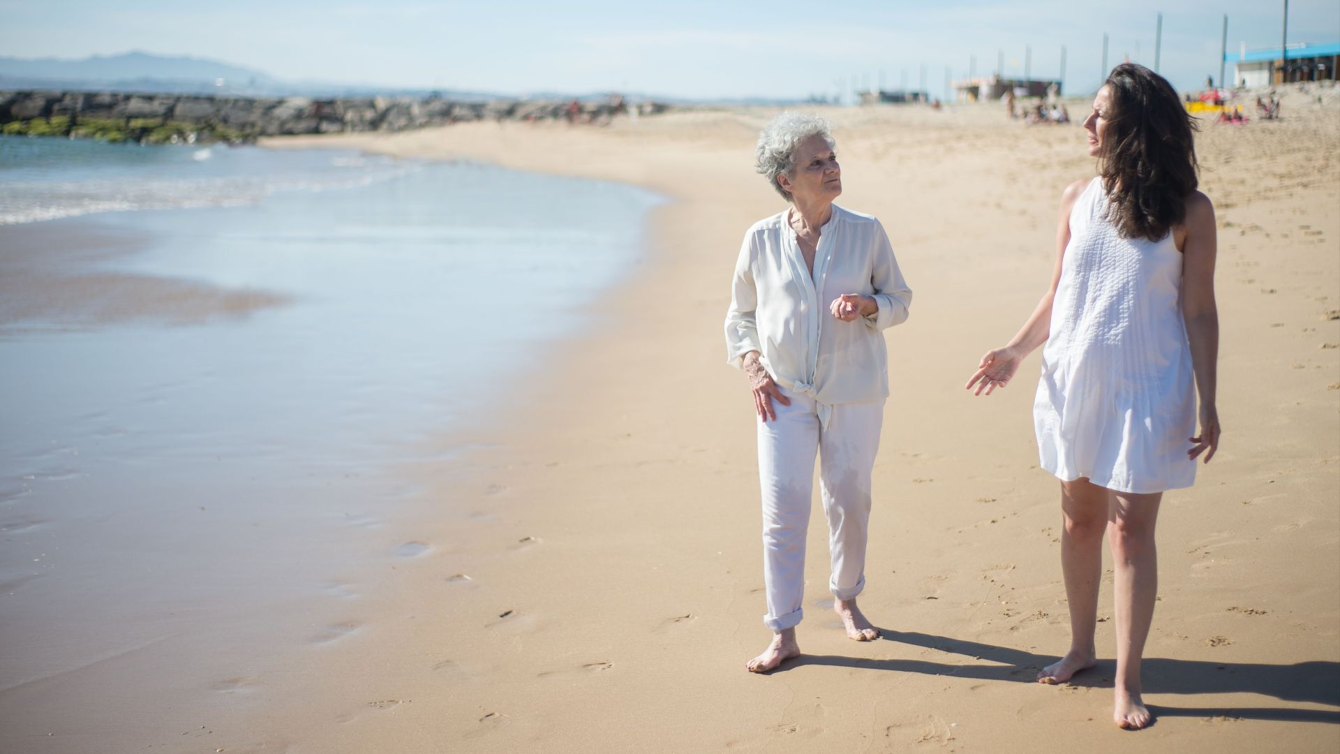 Two women walk barefoot along a sunny beach, enjoying their time together.